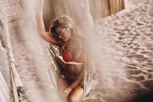curtindo suas férias. vista superior de uma jovem atraente em trajes de banho sorrindo enquanto está sentado na rede na praia foto