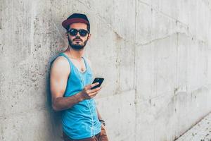 curtindo seu estilo de rua. jovem bonito em fones de ouvido segurando o telefone inteligente e desviar o olhar em pé ao ar livre e encostado na parede de concreto foto