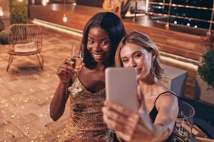 duas mulheres bonitas em vestidos de noite fazendo selfie e sorrindo enquanto passam o tempo na festa de luxo