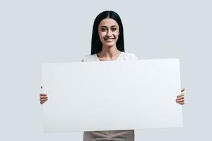 lindo e cheio de energia. jovem mulher confiante segurando flipchart em branco e olhando para a câmera em pé contra um fundo cinza foto