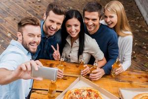 sorriso feliz para selfie vista superior do grupo de jovens segurando garrafas com cerveja e fazendo selfie no telefone inteligente em pé ao ar livre foto