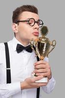 beijando seu troféu. jovem de gravata borboleta segurando um troféu e beijando-o em pé contra um fundo cinza foto