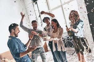 melhor aniversário. jovem feliz comemorando aniversário entre amigos em pé na sala com confetes voando ao redor foto