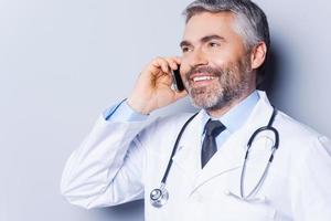 boas notícias. médico de cabelos grisalhos maduros confiante falando no celular e sorrindo em pé contra um fundo cinza foto