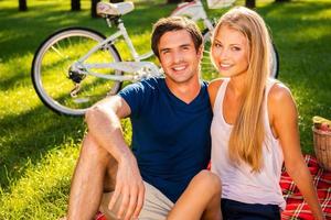um bom dia para piquenique. feliz casal apaixonado relaxando no parque juntos enquanto está sentado na manta de piquenique e sorrindo enquanto bicicleta em pé no fundo foto