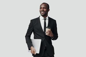 feliz homem africano em trajes formais olhando para longe e sorrindo em pé contra um fundo cinza foto