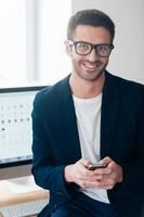 inteligente e confiante. jovem confiante segurando o telefone inteligente e sorrindo enquanto se inclina na mesa do escritório foto