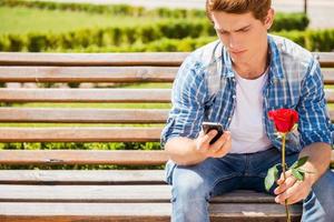 esperando sua namorada. jovem preocupado segurando uma única rosa e olhando para o celular enquanto está sentado no banco foto