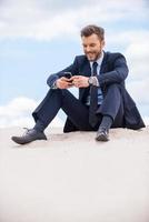 mantendo contato. alegre jovem empresário olhando para seu celular e sorrindo enquanto está sentado na areia e contra o céu azul foto