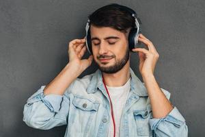 sua música favorita. jovem ajustando seus fones de ouvido e mantendo os olhos fechados em pé contra um fundo cinza foto