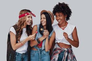 três mulheres jovens atraentes comendo pirulito e sorrindo em pé contra um fundo cinza foto