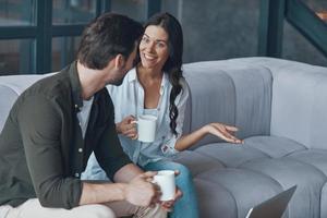 lindo casal jovem tomando café e se comunicando enquanto está sentado no sofá em casa foto