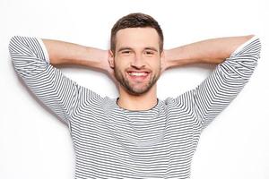 sentindo-se calmo e satisfeito. jovem bonito segurando as mãos atrás da cabeça e sorrindo em pé contra um fundo branco foto