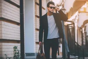 sempre em grande estilo. jovem bonito em roupa casual inteligente carregando bolsa e ajustando seus óculos de sol em pé na rua foto