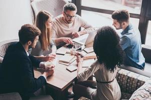 preste atenção a este jovem bonito apontando para tablet digital enquanto está sentado na mesa do escritório na reunião de negócios com seus colegas de trabalho foto