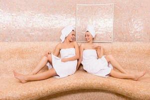 meninas na sauna. duas mulheres jovens atraentes embrulhadas em toalha conversando e sorrindo enquanto passam o tempo na sauna foto