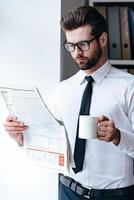 lendo jornal fresco. pensativo jovem empresário em óculos lendo jornal e segurando a xícara de café em pé no escritório foto