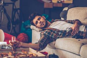 jovem bonito segurando uma fatia de pizza e lata de cerveja enquanto estava deitado no sofá na sala bagunçada depois da festa foto