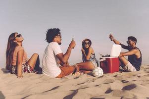 passar bons momentos com os amigos. vista traseira de quatro jovens alegres passando um bom tempo juntos enquanto estão sentados na praia e bebendo cerveja foto