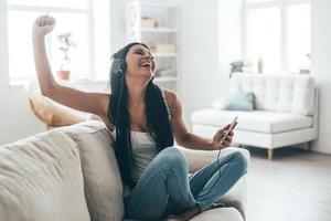 desfrutando de música favorita jovem feliz em fones de ouvido segurando o telefone inteligente e gesticulando enquanto está sentado no sofá em casa foto