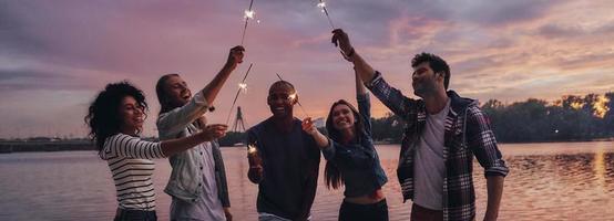 criando memórias felizes. comprimento total de jovens em roupas casuais sorrindo e segurando faíscas em pé no cais foto