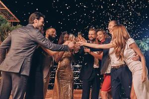 grupo de pessoas bonitas em trajes formais brindando com champanhe e sorrindo enquanto confetes voando ao redor deles foto