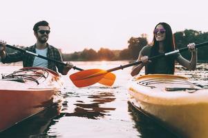 a vida é melhor no rio. alegre casal jovem andando de caiaque no rio junto com o pôr do sol ao fundo foto