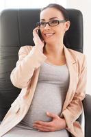 mulher de negócios grávida feliz. linda empresária grávida falando ao telefone e sorrindo enquanto está sentado em seu local de trabalho no escritório foto