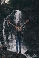 vista traseira de comprimento total de jovem com mochila, mantendo as mãos levantadas em pé perto da cachoeira foto