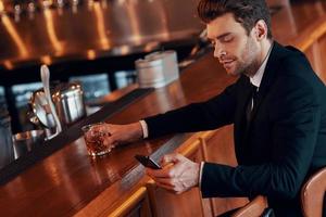 jovem bonito de terno completo usando telefone inteligente enquanto está sentado no balcão do bar no restaurante foto