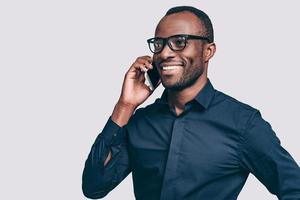 Boa conversa. bonito jovem africano falando no telefone inteligente e sorrindo em pé contra um fundo cinza foto