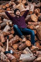 relaxante após um dia de trabalho duro. comprimento total de guarda florestal jovem confiante segurando a cabeça nas mãos e olhando para cima enquanto se inclina para os logs foto