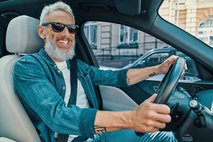 feliz homem sênior em roupas casuais, desfrutando de passeio de carro enquanto está sentado no banco da frente do carro foto