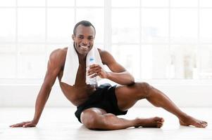 mantendo seu corpo em forma. alegre jovem africano sentado no chão e segurando a garrafa com água foto