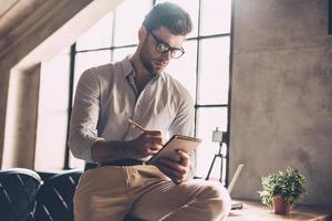 apenas inspirado. jovem confiante em roupas casuais inteligentes, escrevendo algo em seu caderno enquanto se inclina na mesa no escritório foto