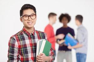 inteligente e confiante. alegre adolescente chinês em óculos segurando livros e sorrindo enquanto seus amigos em pé no fundo foto