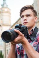 fotógrafo confiante. jovem bonito segurando a câmera digital e desviar o olhar em pé ao ar livre foto