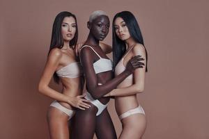 beleza sem esforço. três mulheres atraentes de raça mista, olhando para a câmera em pé contra um fundo marrom foto