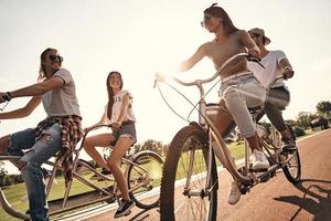 amigos fazem um ao outro feliz. grupo de jovens felizes em roupas casuais sorrindo enquanto andam de bicicleta juntos ao ar livre foto