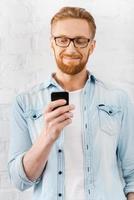 sempre conectado. feliz jovem barbudo segurando o celular e sorrindo em pé contra a parede de tijolos foto