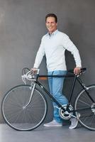 homem com bicicleta. alegre homem maduro em pé perto de sua bicicleta com fundo cinza foto