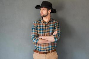 cowboy sério e confiante. jovem bonito com chapéu de cowboy, mantendo os braços cruzados e olhando para longe em pé contra um fundo cinza foto