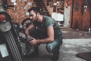 o que parece ser um problema jovem confiante segurando um pano e olhando para a motocicleta enquanto se agacha perto dela com garagem ao fundo foto