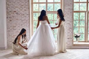 início do dia mais feliz. comprimento total de damas de honra ajudando a noiva com seu vestido de casamento em pé perto da janela juntos foto