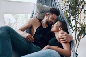 ele nunca vai deixá-la ir. lindo casal jovem abraçando e sorrindo enquanto descansava na rede dentro de casa foto