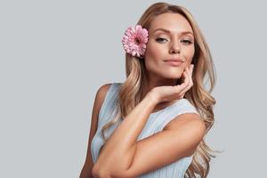 verdadeira beleza feminina. mulher jovem e atraente com uma flor no cabelo olhando para a câmera e sorrindo em pé contra um fundo cinza foto