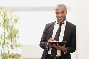 empresário com bloco de notas. alegre jovem empresário africano escrevendo algo em seu bloco de notas e sorrindo para a câmera foto