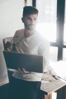 pensando em solução. jovem pensativo carregando laptop e óculos enquanto se inclina na mesa no escritório e desvia o olhar foto