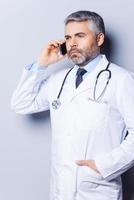 médico ao telefone. médico de cabelos grisalhos maduros confiante falando no celular e desviando o olhar em pé contra um fundo cinza foto