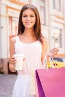 linda viciada em compras. bela jovem sorridente segurando sacolas de compras e copo de bebida quente em pé ao ar livre foto